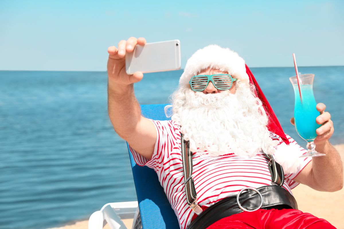 Santa Claus on the beach holding a cold drink and taking a selfie for Christmas brunch in June