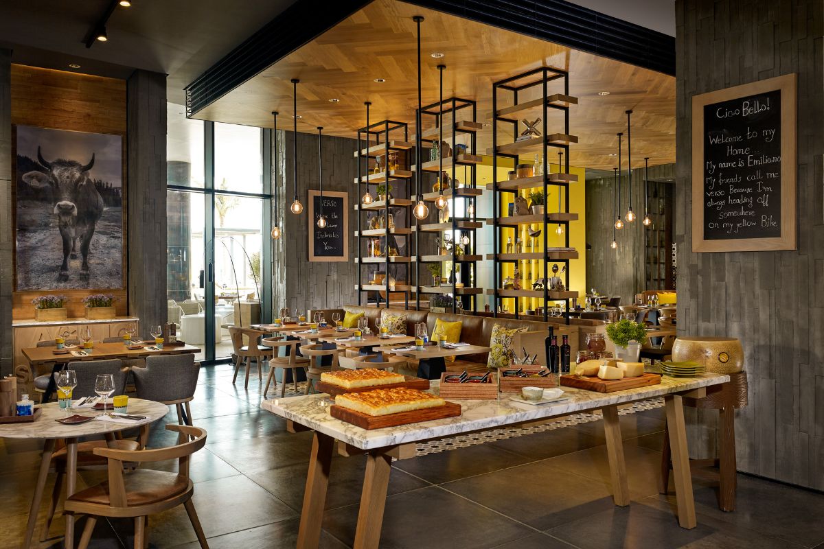 Verso interior with tables set up, a picture of a cow framed on the wall, a menu board with writings on them, foccacia bread on the table together with some cheese like grana padano