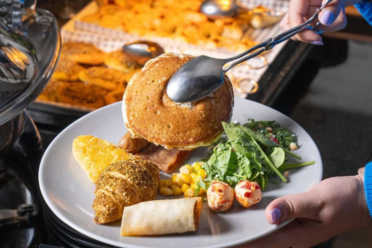 Cafe 302 at Al Maha Arjaan by Rotana breakfast buffet for the best breakfast offers in Abu Dhabi, pictured a lady holding a tong with a plate full of breakfast items such as a pancake, zaatar croissant, egg, salad and more