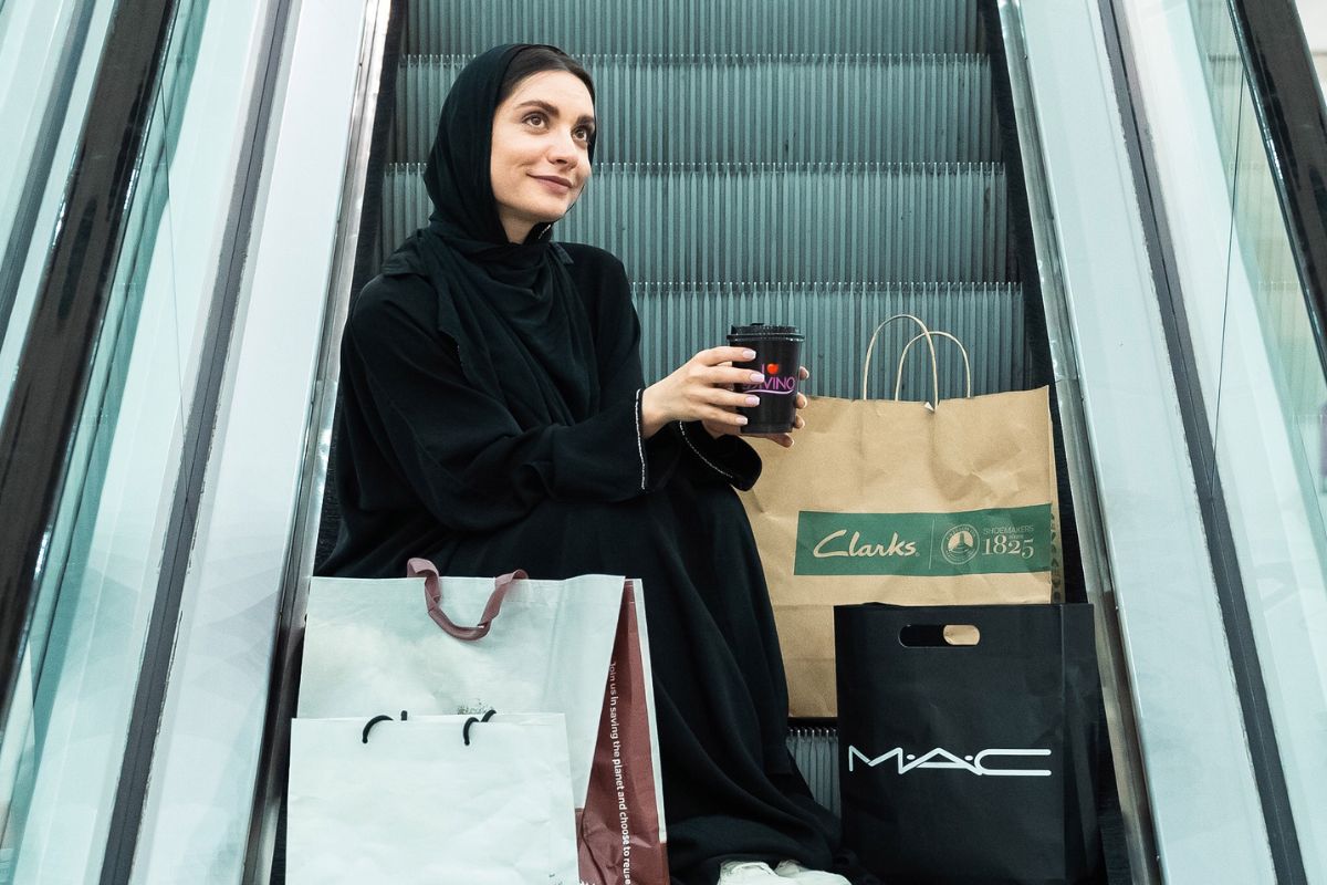 An Emirati woman sitting on an escalator with shopping bags and a coffee cup at Al Ain Mall shopping center