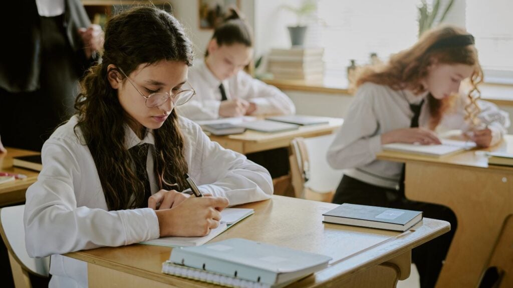 Three students are studying in a classroom setting. The students are seated at wooden desks and writing in notebooks.