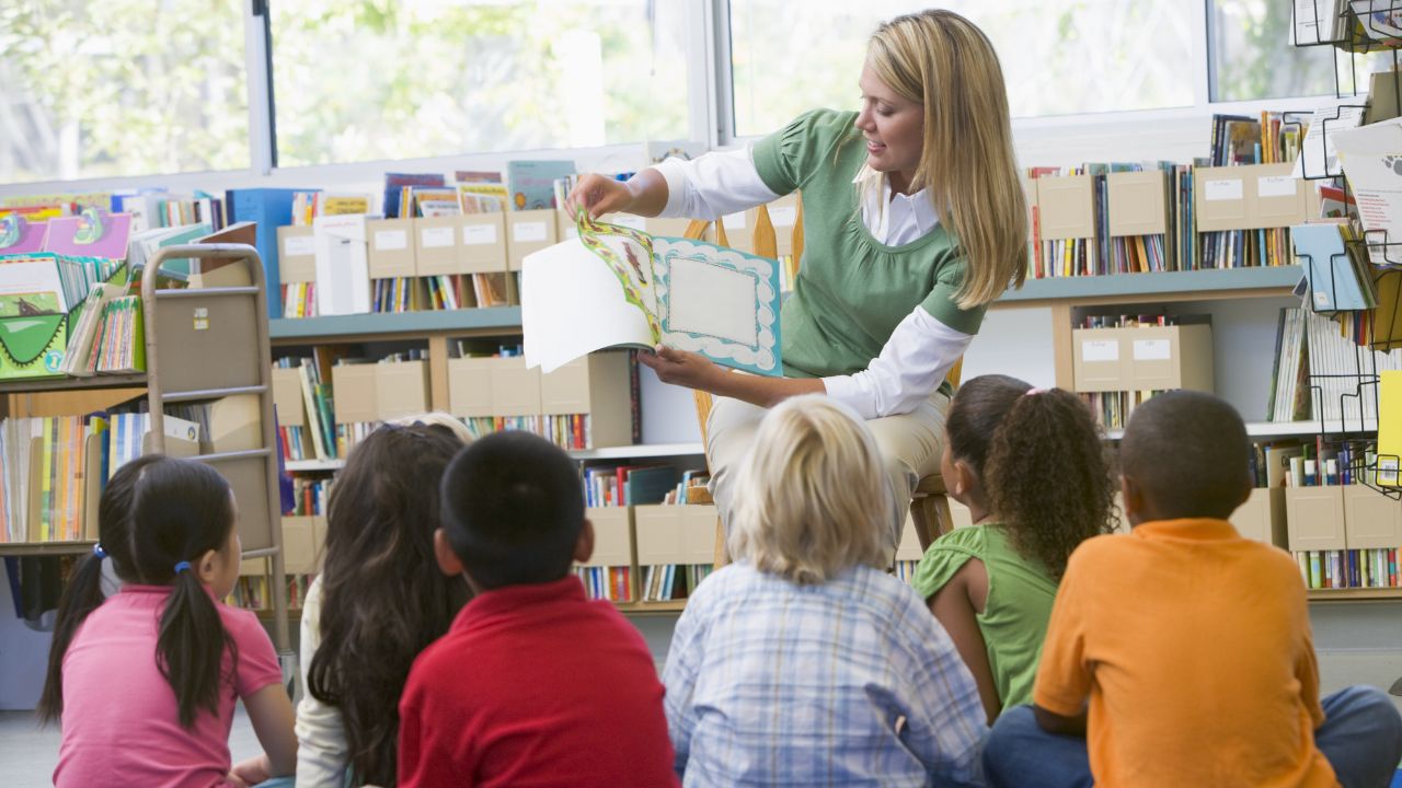 female teacher reading a book to her young students sitting down